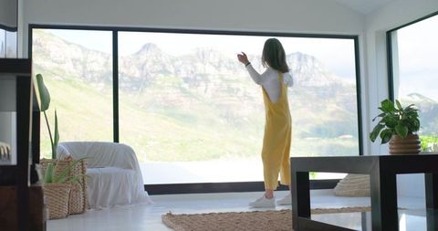 Woman in yellow dress gazing through panoramic window at mountain view, minimal livingroom