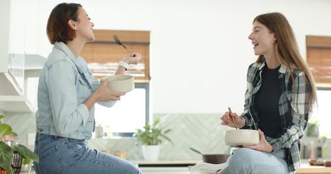 Two women chatting and eating from bowls on bright kitchen island, casual friendship