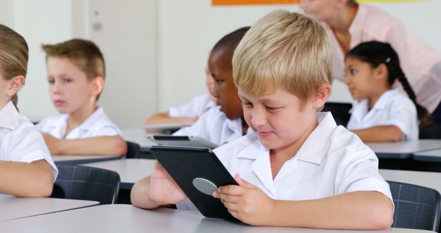 Group of young children wearing white uniforms using tablets in a busy classroom emphasizes modern education with technology. Ideal for websites and articles focused on education, technology in education, primary school environments, and interactive learning methods. Suitable for educational blogs, school promotional materials, and e-learning resources.