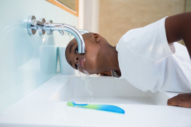 Side view of boy washing mouth from faucet over sink at bathroom
