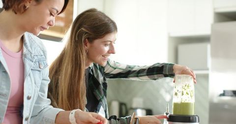 Two diverse young women preparing green smoothie while chopping vegetables in kitchen