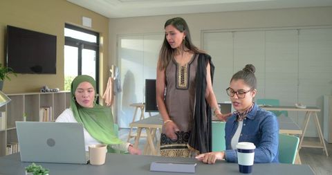 Multicultural women collaborating in office wearing hijab, kurta and denim with laptop and tablet