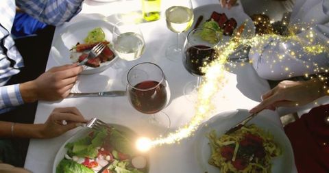 Overhead dinner party with wine glasses, pasta and salad, guests serving amid golden sparkles