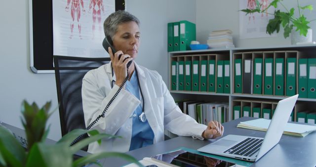 Female doctor, wearing white coat and stethoscope, is talking on phone in medical office while using laptop. Medical charts and folders visible in background. Ideal for depicting healthcare, medical consultancy, senior professionals, and communication in medical settings.