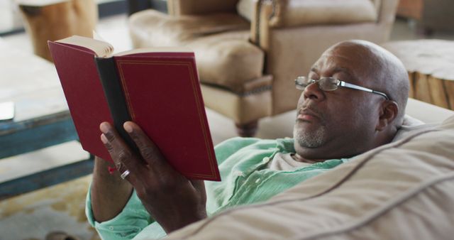 A mature man with glasses is comfortably relaxing on a couch while reading a red hardcover book in a cozy living room. This can be ideal for illustrating themes like relaxation, leisure activities, learning, and peaceful moments. Great for representing senior lifestyle, reading habits, and comfort at home.