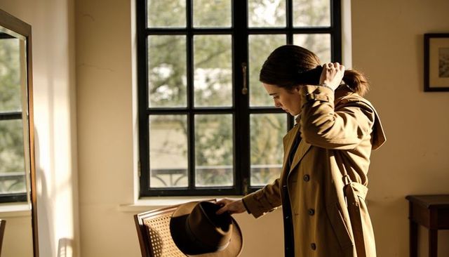 Woman adjusting hair in trench coat holding hat by sunlit window elegant lifestyle