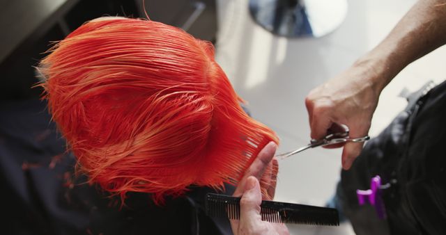 A professional hairdresser trims bright orange hair with precision scissors. This scene captures a close-up of the hands, scissors, and comb in action within a salon setting. This image is ideal for use in content related to hair styling, beauty services, or professional grooming tutorials, emphasizing creativity and professional techniques.