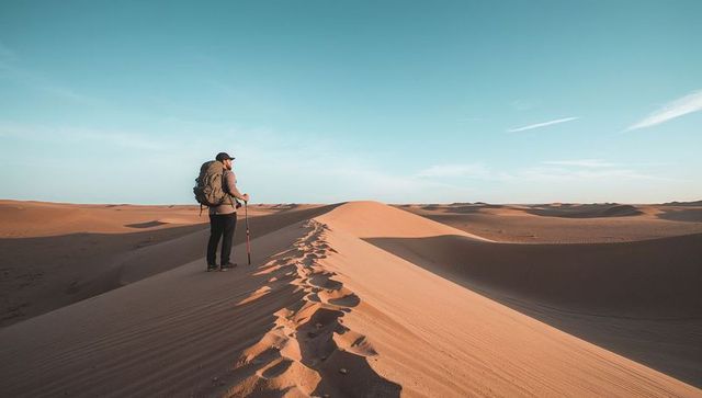 Hiker standing on desert dune crest at golden hour with backpack and trekking pole