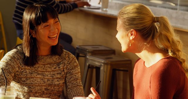 The photograph shows two women smiling and engaging in conversation at a cafeteria table. Both have drinks in front of them, and the atmosphere suggests they are enjoying a friendly chat during the daytime. This image can be used for promoting social interactions, friendship, and casual meet-ups in coffee shops or eatery environments.