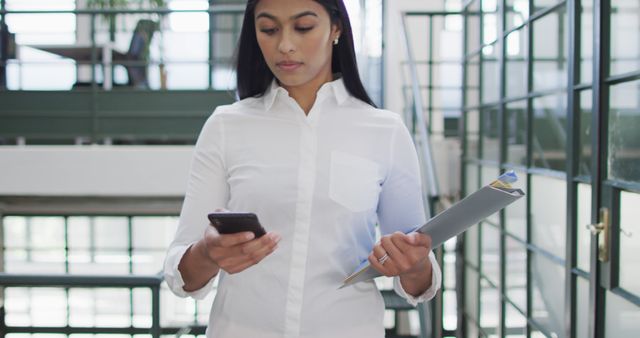 Businesswoman in white shirt using smartphone and holding documents in modern office. Ideal for business, corporate presentations, brochures, advertisements, and articles related to workplace productivity, modern work environments, and professional life.