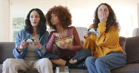 Three Women Playing Video Games and Sharing Chips, Laughing and Reacting on Cozy Living Room Couch