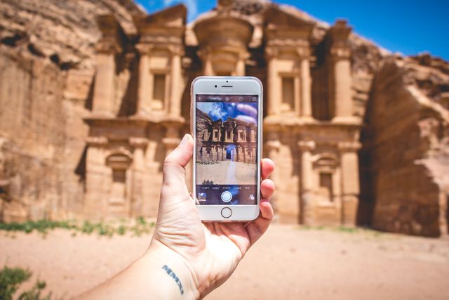 Hand holding smartphone capturing a photo of the ancient Petra site emphasizes the mix of modern technology with historic tourism. Useful for travel agencies, technology firms, and cultural blogs. Highlights intersection of old and new, appealing to adventure seekers and history enthusiasts.