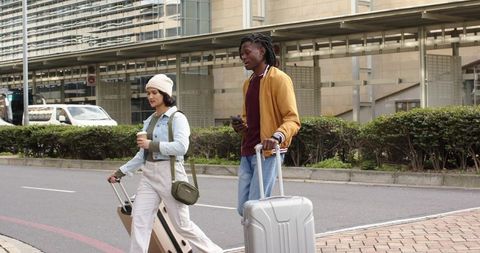 Diverse travelers walking curbside pulling suitcases outside modern airport terminal