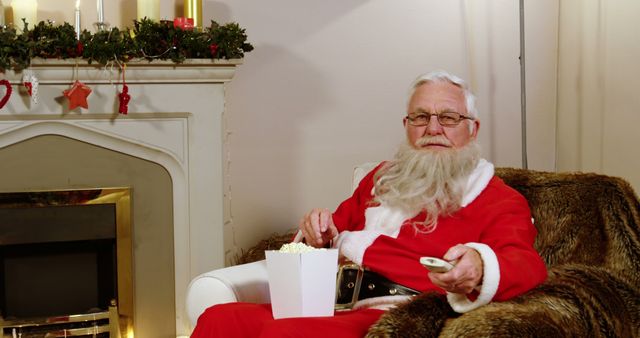 A Caucasian senior man dressed as Santa Claus is relaxing on a chair with popcorn and a remote control, with copy space. His leisurely pose in a festive room suggests a break from holiday activities.