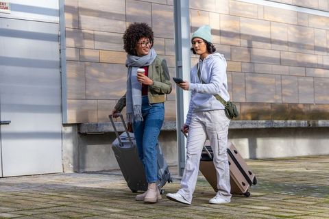 Two friends walking with suitcases in urban plaza checking phone holding coffee