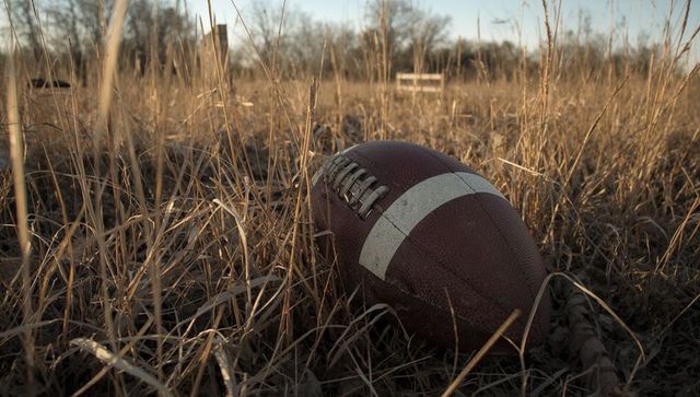 Weathered football nestling in dry grass at golden hour with empty bench and bare trees
