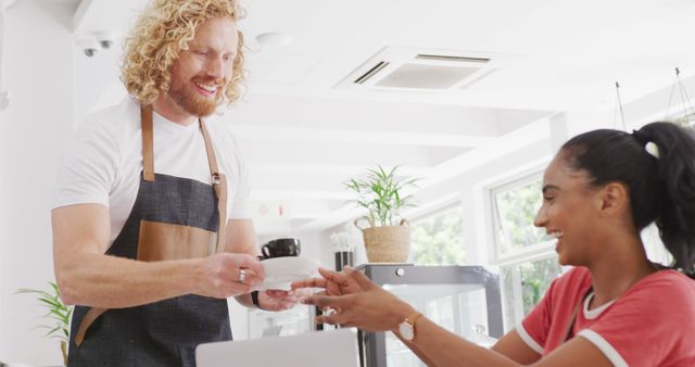 A male barista with curly blonde hair is serving coffee to a smiling female customer in a modern café with a clean and bright interior. This image is perfect for marketing materials related to hospitality, customer service, or coffee culture. It portrays a warm and welcoming atmosphere, ideal for use in websites, brochures, and ads promoting cafés, barista courses, or customer satisfaction.