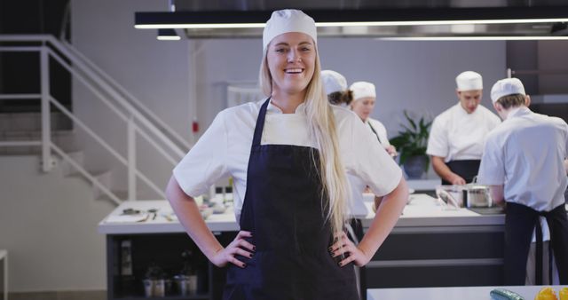 Confident female chef standing in modern professional kitchen, smiling and posing with hands on her hips. Additional chefs in the background working on culinary tasks. Suitable for promoting culinary schools, chef training programs, cooking classes, and professional kitchen environments.