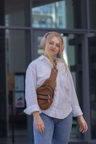 Young woman listening to music with pink headphones wearing brown crossbody bag in urban lobby