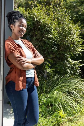 Leaning at doorway smiling woman wearing rust shirt and jeans in sunny garden