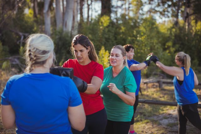 Women engaging in boxing training in an outdoor boot camp, surrounded by nature on a sunny day. Ideal for promoting fitness programs, outdoor activities, group exercise classes, and healthy lifestyle campaigns. Suitable for use in fitness blogs, sports advertisements, and wellness websites.