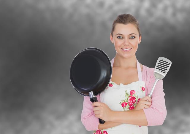 Woman holding frying pan and skimmer, wearing floral apron. Excellent for content related to cooking, culinary blogs, recipe websites, or kitchen utensil advertisements. Highlights kitchen preparation and cooking activities.