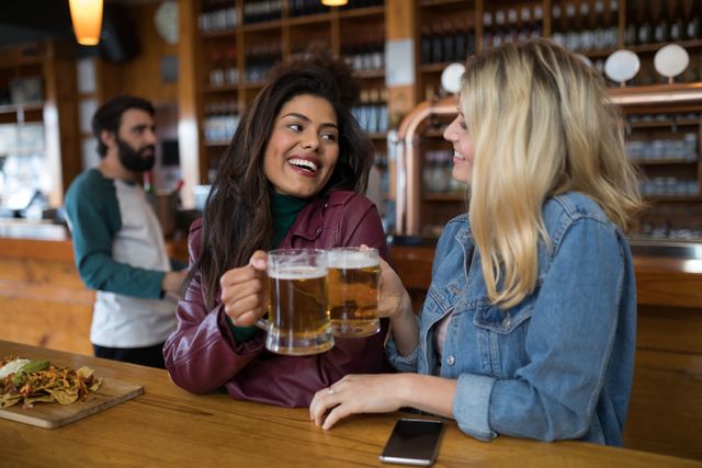 Smiling female friends toasting glass of beer at counter in bar
