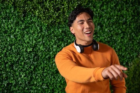 Asian man laughing and pointing wearing orange sweatshirt and headphones against green wall