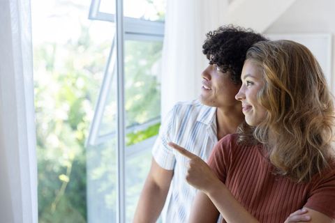 Couple enjoying morning light pointing at garden through open window, home harmony