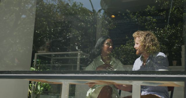 Two people chatting and enjoying coffee inside a cozy coffee shop while sitting by a large window with green plants and sunlight filtering through. Could be used for promoting cafés, coffee brands, relaxation or representing casual conversations and bonding moments.