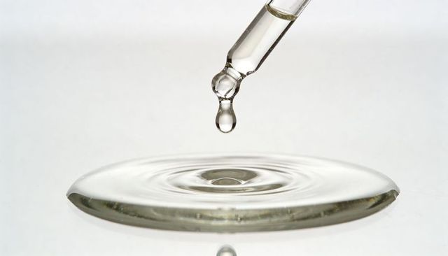 Glass dropper dripping clear serum above rippled liquid surface minimalist sterile high-key