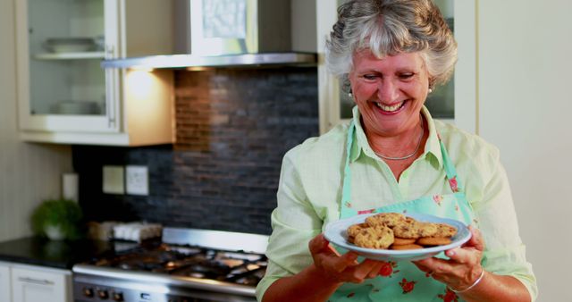 Senior woman holding a plate of freshly baked cookies in a modern kitchen. She is smiling and looks delighted with her baking. This image is perfect for use in advertisements or websites related to home baking, cooking classes, culinary arts, family recipes, or kitchen appliances. Also suitable for illustrating stories, articles, and blog posts about enjoying the simple pleasures of home-cooked food, sharing family traditions, or senior lifestyles.