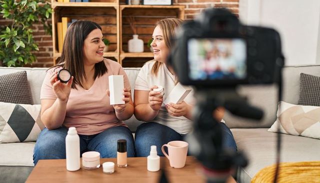 Smiling women filming makeup and skincare tutorial on sofa, demonstrating beauty products