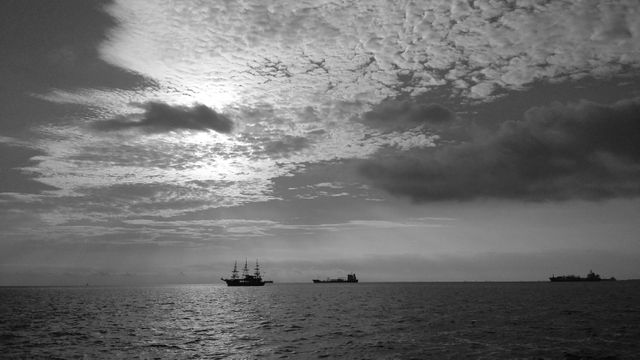 Ideal for conveying a sense of tranquility and adventure, this black and white image of sailing ships silhouetted against a dramatic sunset sky can be used in travel blogs, maritime publications, or as a decorative poster.