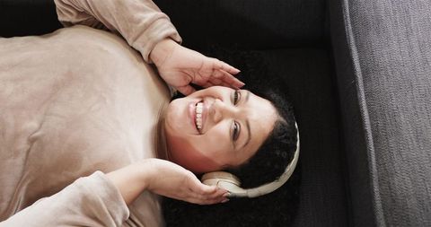 Smiling woman relaxing on sofa wearing headphones and cozy beige top listening to music