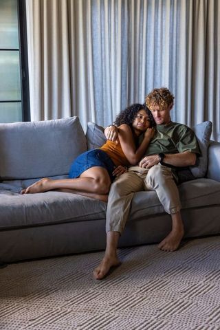 Diverse couple cuddling and relaxing on light-gray sofa, cozy at-home intimacy
