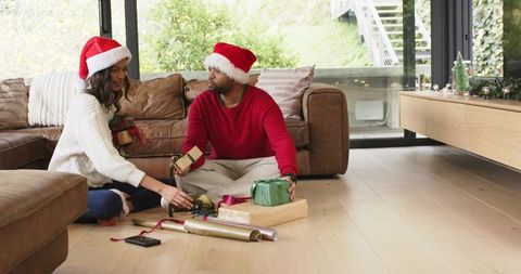 Diverse couple wearing santa hats sitting on living room floor wrapping christmas gifts