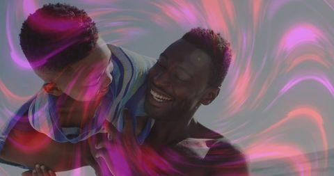 Father lifting son among vibrant pink light patterns at beach