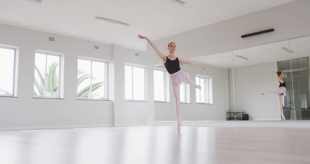 Ballet dancer practicing in a bright studio with large windows and mirrors, wearing a black leotard and white tutu, standing with one leg raised. Ideal for illustrating professional dance training, performing arts, elegance in motion, artistic expression, fitness activities, dance classes, and disciplined practice.