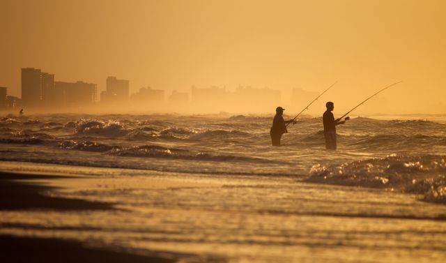 Silhouetted fishermen casting lines into ocean at sunset with distant city buildings. Captured at dusk, against glowing sky, with rolling waves. Ideal for themes on outdoor recreation, seaside activities, serene moments, and nature appreciation. Suitable for travel blogs, leisure advertising, and backgrounds.