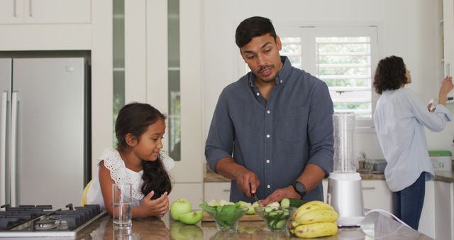 Father and daughter are preparing a healthy smoothie together in a modern kitchen. The father is cutting fruits while his daughter watches and assists. A blender and various fruits such as bananas and apples are on the counter. This can be used for promoting family bonding, healthy lifestyle, cooking activities, and parenting moments.