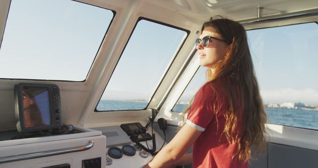 Young woman steering a boat on a sunny day, wearing sunglasses. Ideal for depicting freedom, adventure, travel, and nautical activities. Can be used in advertisements for travel agencies, boating equipment, or lifestyle articles that focus on outdoor adventures and recreational water activities.