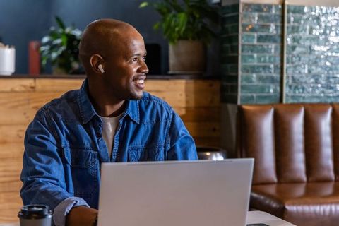 African american man smiling and working on laptop in cozy cafe wearing earbuds