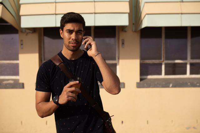 Young biracial man standing outdoors on a sunny day, talking on his smartphone while holding a coffee. Ideal for use in advertisements, blogs, or articles about modern communication, technology, urban lifestyle, or leisure activities.