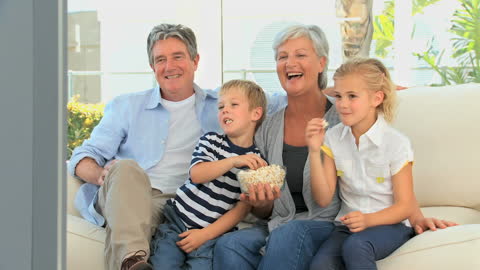 Grandparents Watching TV with Grandchildren on Sofa, Smiling and Eating Popcorn
