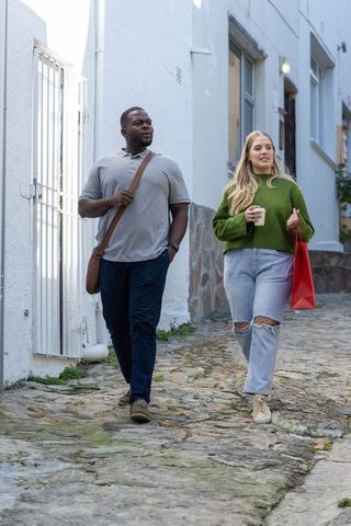 Diverse couple walking down narrow cobblestone street carrying coffee and red shopping bag