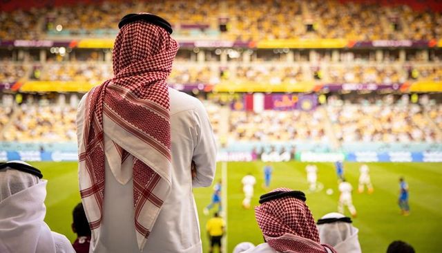 Gulf spectator wearing traditional thobe and red shemagh watching live football match in stadium