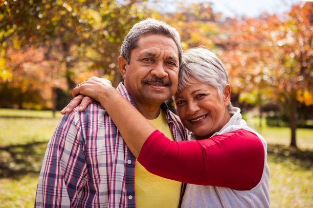 Elderly couple enjoying a warm embrace in a park during autumn. The vibrant fall foliage in the background adds a touch of color and warmth to the scene. Ideal for use in advertisements, articles, or brochures related to senior living, retirement, family values, and outdoor activities for seniors.