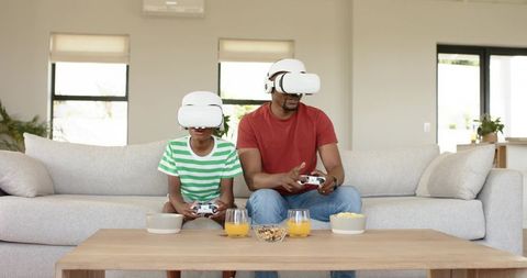 Father and Son Playing Virtual Reality Games on Sofa with Controllers and Snacks