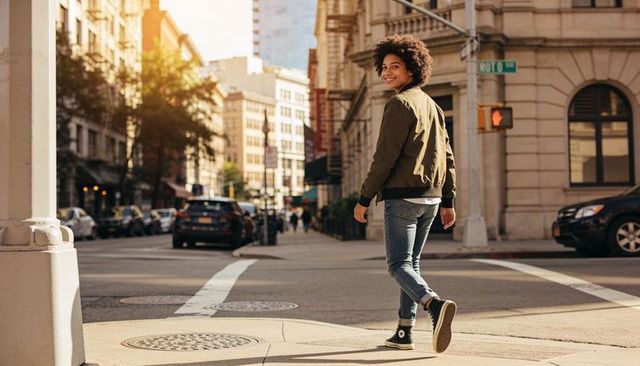 Smiling woman walking back across urban crosswalk during golden hour in olive bomber jacket